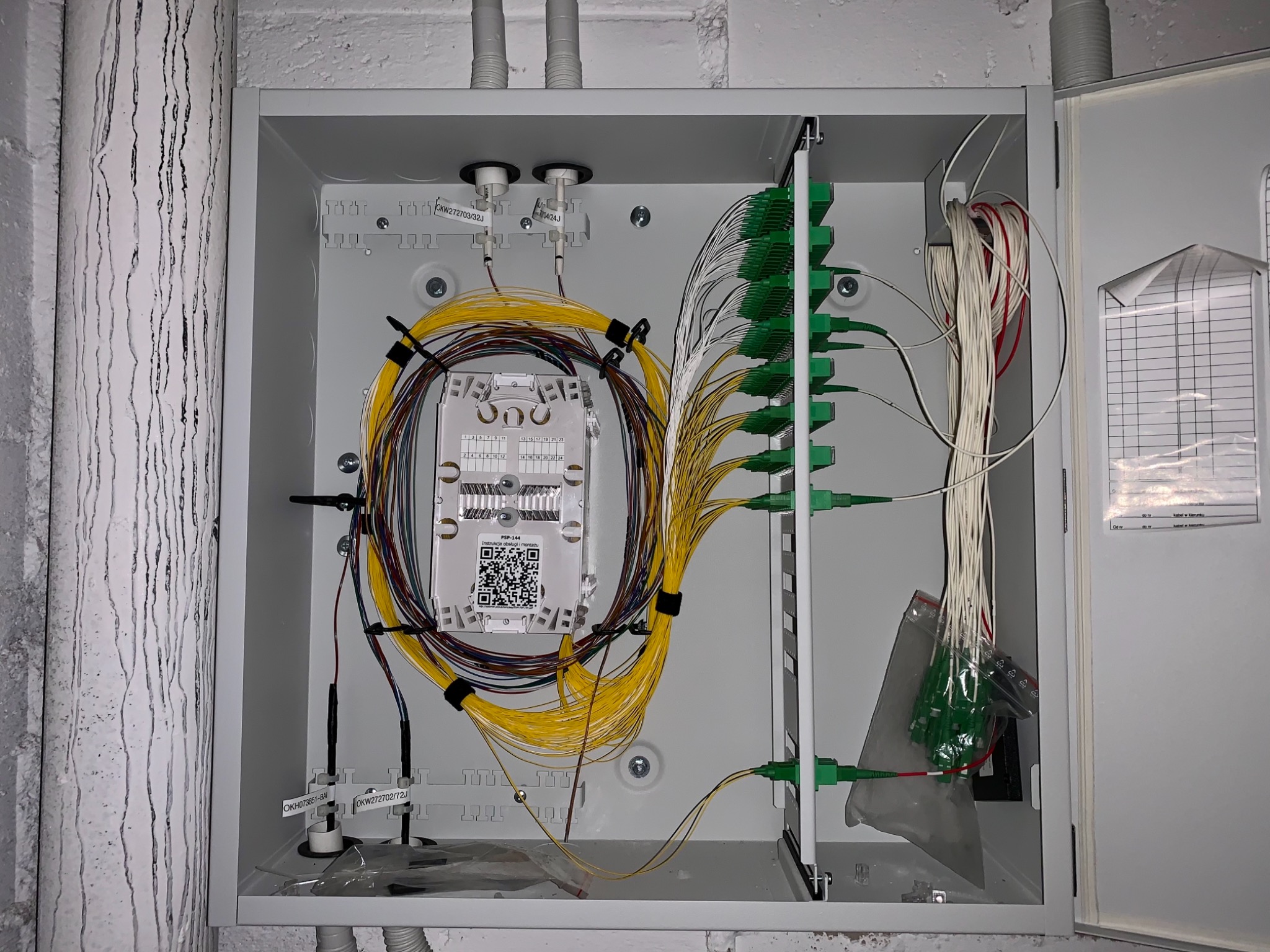 Otwarta skrzynka telekomunikacyjna z rozdzielnicą światłowodową, widoczne kable światłowodowe w kolorze żółtym, zielone złącza, etykiety identyfikacyjne kabli i QR kod na module. Skrzynka...