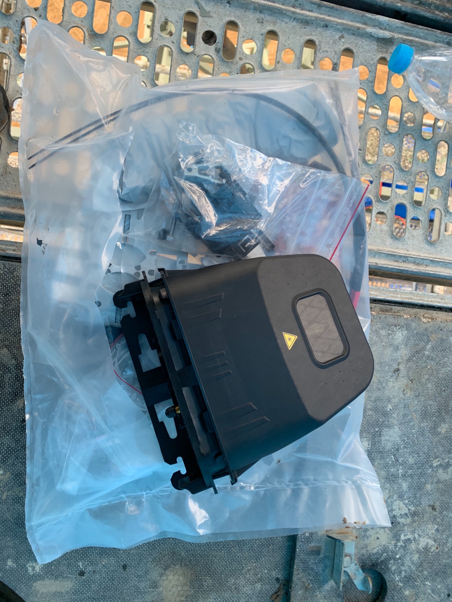 Elementy instalacji telekomunikacyjnej w plastikowej torbie, widoczna czarna obudowa z żółtym symbolem trójkąta i inne akcesoria, całość leży na metalowej kratownicy.