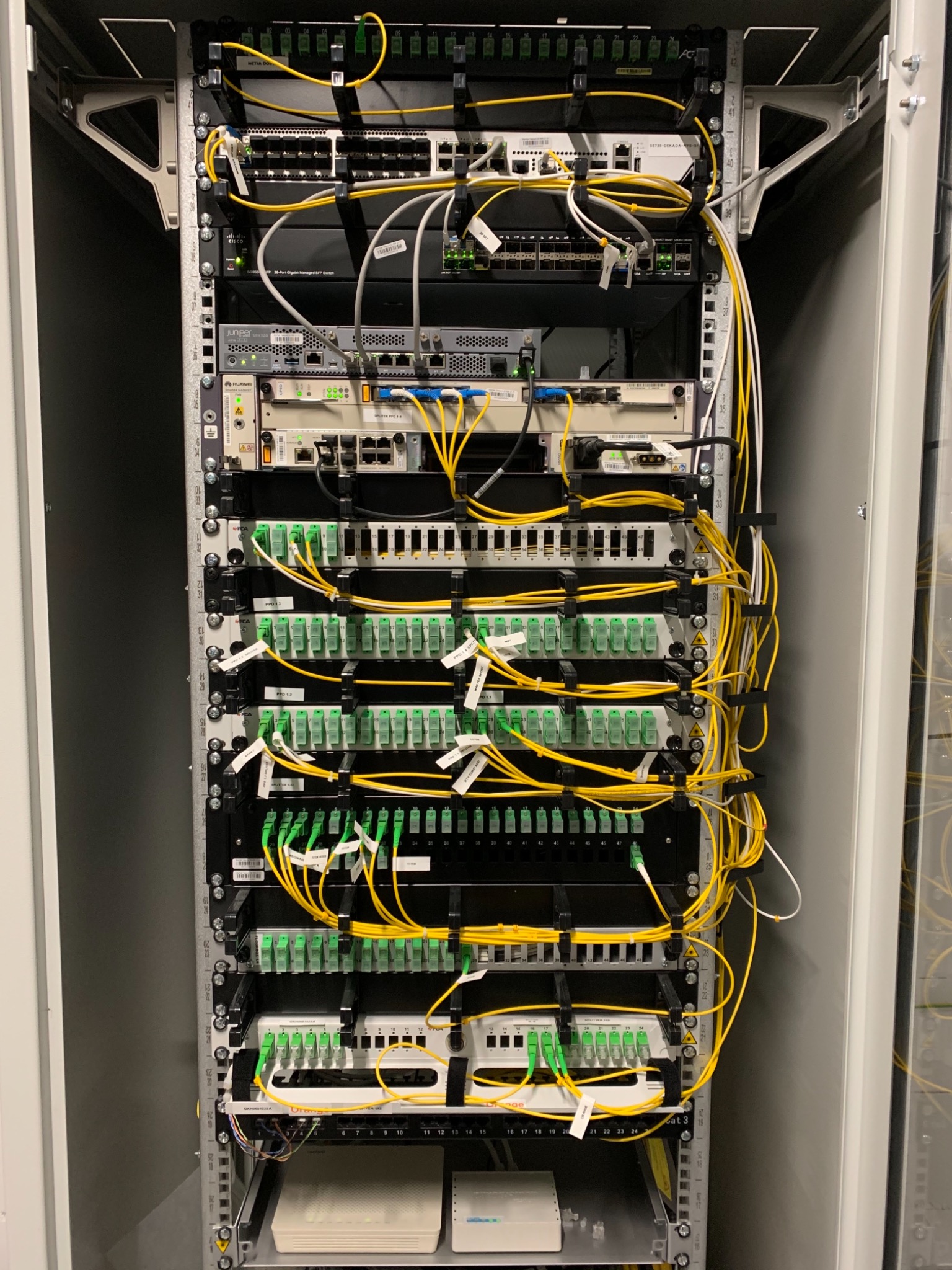 Szafa rack z panelami światłowodowymi, przełącznikami sieciowymi i okablowaniem w kolorze żółtym i zielonym. Widoczne urządzenia marek Huawei, Juniper i Cisco. Elementy zamontowane w metalowej...