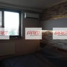 Klimatyzator ścienny zamontowany w sypialni z dekoracyjną ścianą z kamienia i grzejnikiem pod oknem z żaluzjami.