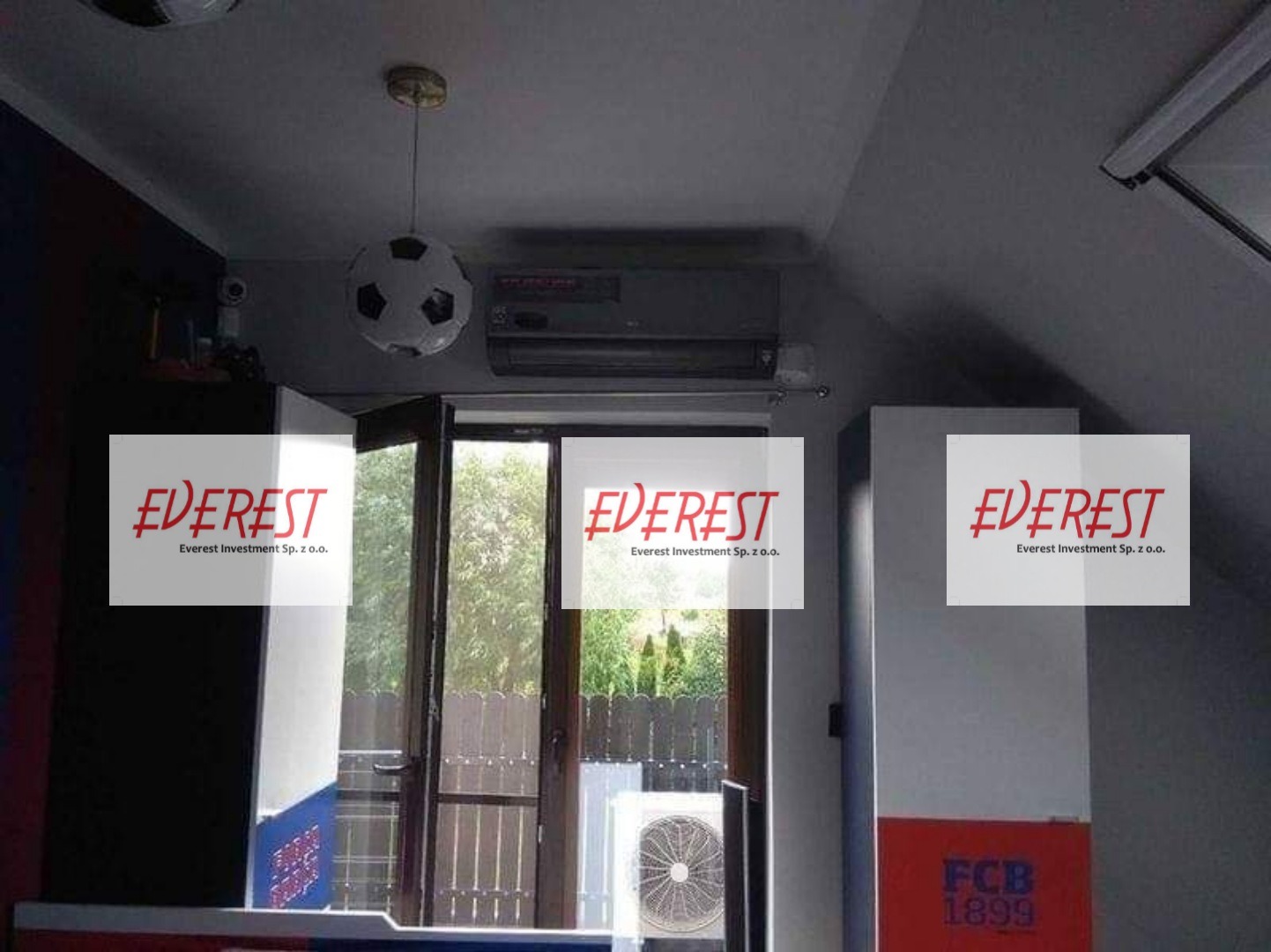 Wnętrze pokoju z zamontowanym klimatyzatorem pod sufitem, lampą w kształcie piłki nożnej, otwartymi drzwiami balkonowymi i jednostką zewnętrzną klimatyzacji widoczną na balkonie.