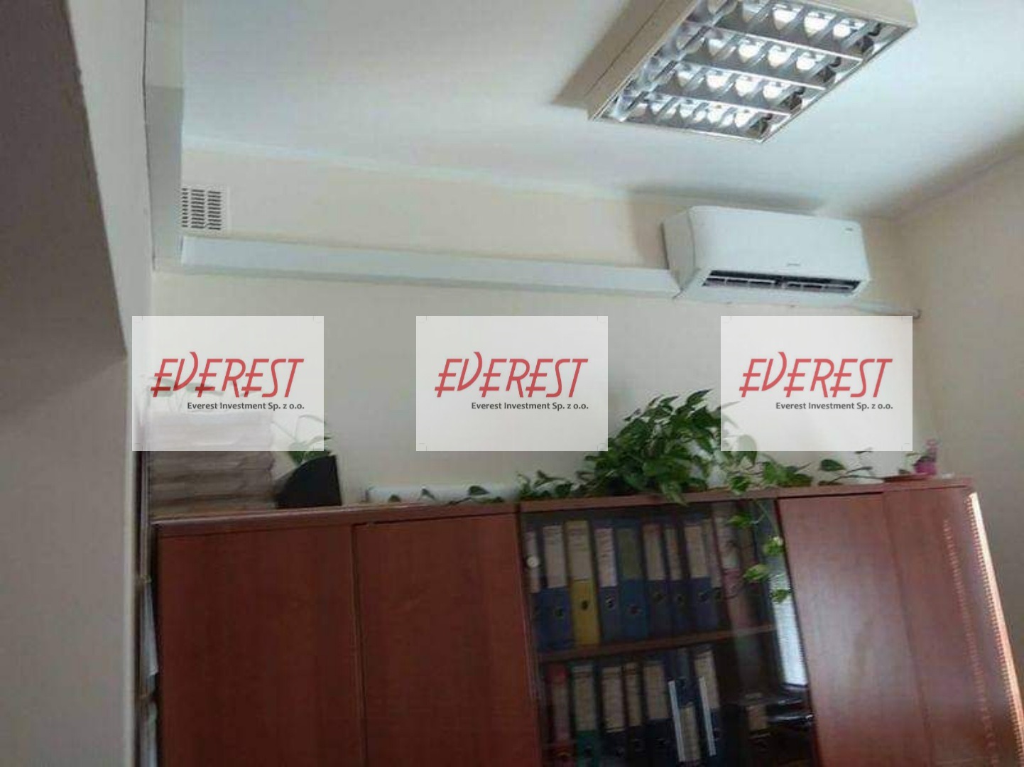 Klimatyzator ścienny zamontowany w biurze nad szafą z segregatorami, widoczna lampa sufitowa i kratka wentylacyjna, na zdjęciu nałożone logo firmy.