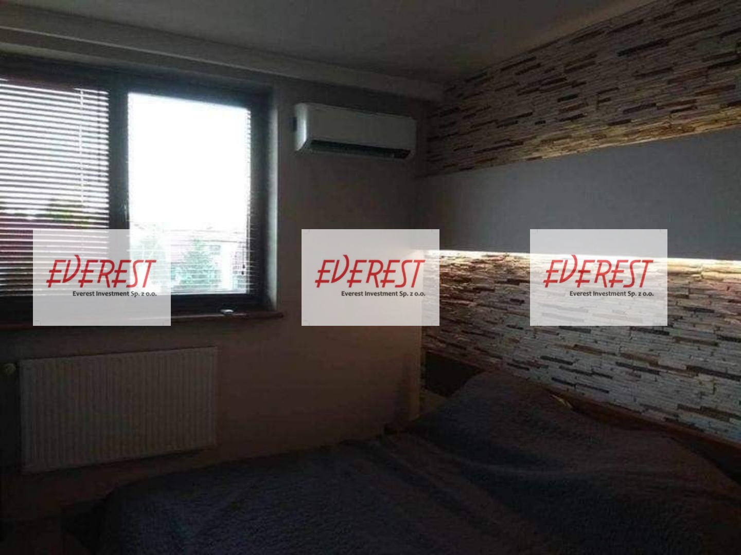 Klimatyzator ścienny zamontowany w sypialni z dekoracyjną ścianą z kamienia i grzejnikiem pod oknem z żaluzjami.