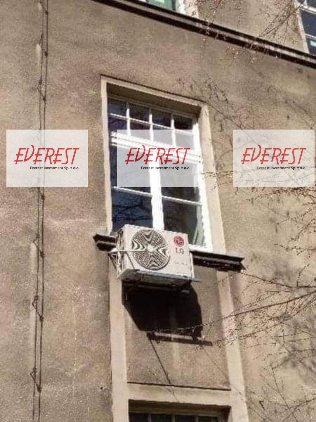 Zewnętrzna jednostka klimatyzacji LG zamontowana pod oknem w starym, szarym budynku z widocznymi śladami zabrudzeń i instalacji elektrycznej na elewacji.