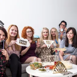 Grupa uśmiechniętych kobiet siedzi na kanapie trzymając rekwizyty z napisami 'Coming Soon', 'Ready to pop', 'Baby Inside', 'Welcome Baby', 'Congrats!', na stole sałatki i przekąski, złote balony...