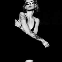 Marcin Frej Photography - Czarno-biały portret kobiety w okularach, z tatuażami na ramionach, na ciemnym tle. Ekspresyjna poza i ostre światło.