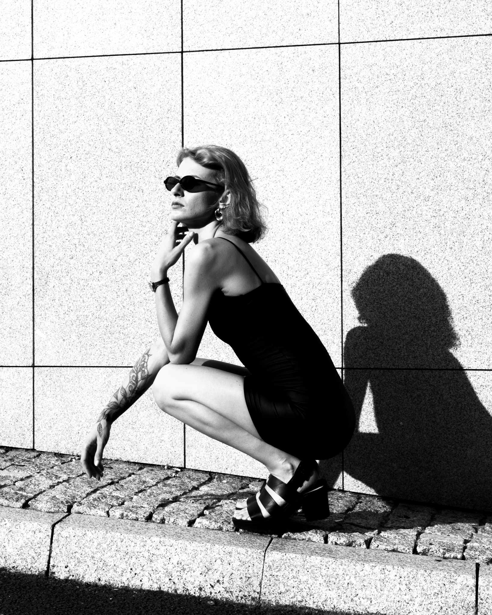 Kobieta w czarnej sukience kuca na tle kamiennej ściany i chodnika, nosi okulary i ma tatuaż na ramieniu. Zdjęcie czarno-białe, ostre światło tworzy cień.