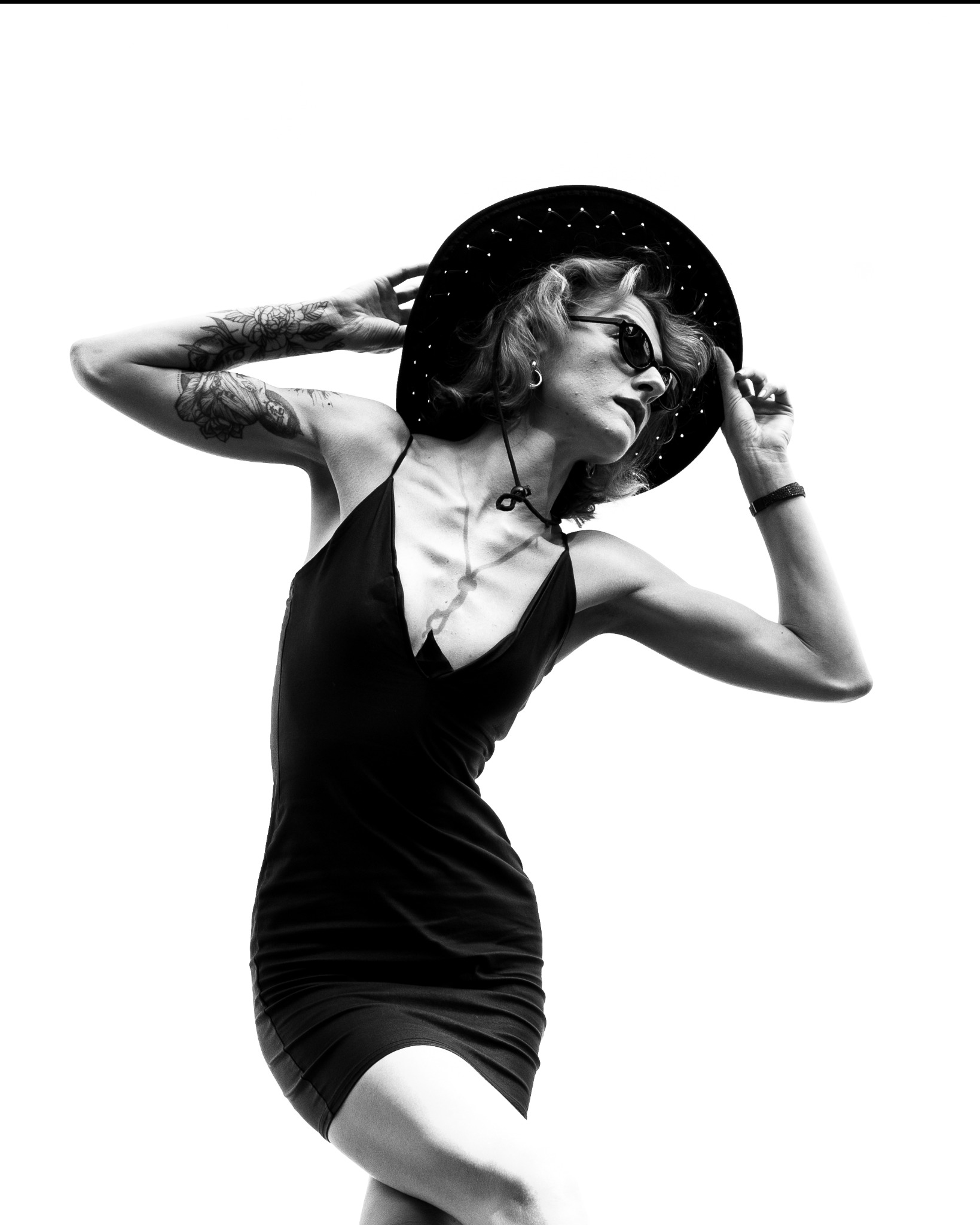 Czarno-białe zdjęcie kobiety w kapeluszu i sukience na jasnym tle, z widocznymi tatuażami na ramionach. Ujęcie z dołu podkreśla sylwetkę modelki.