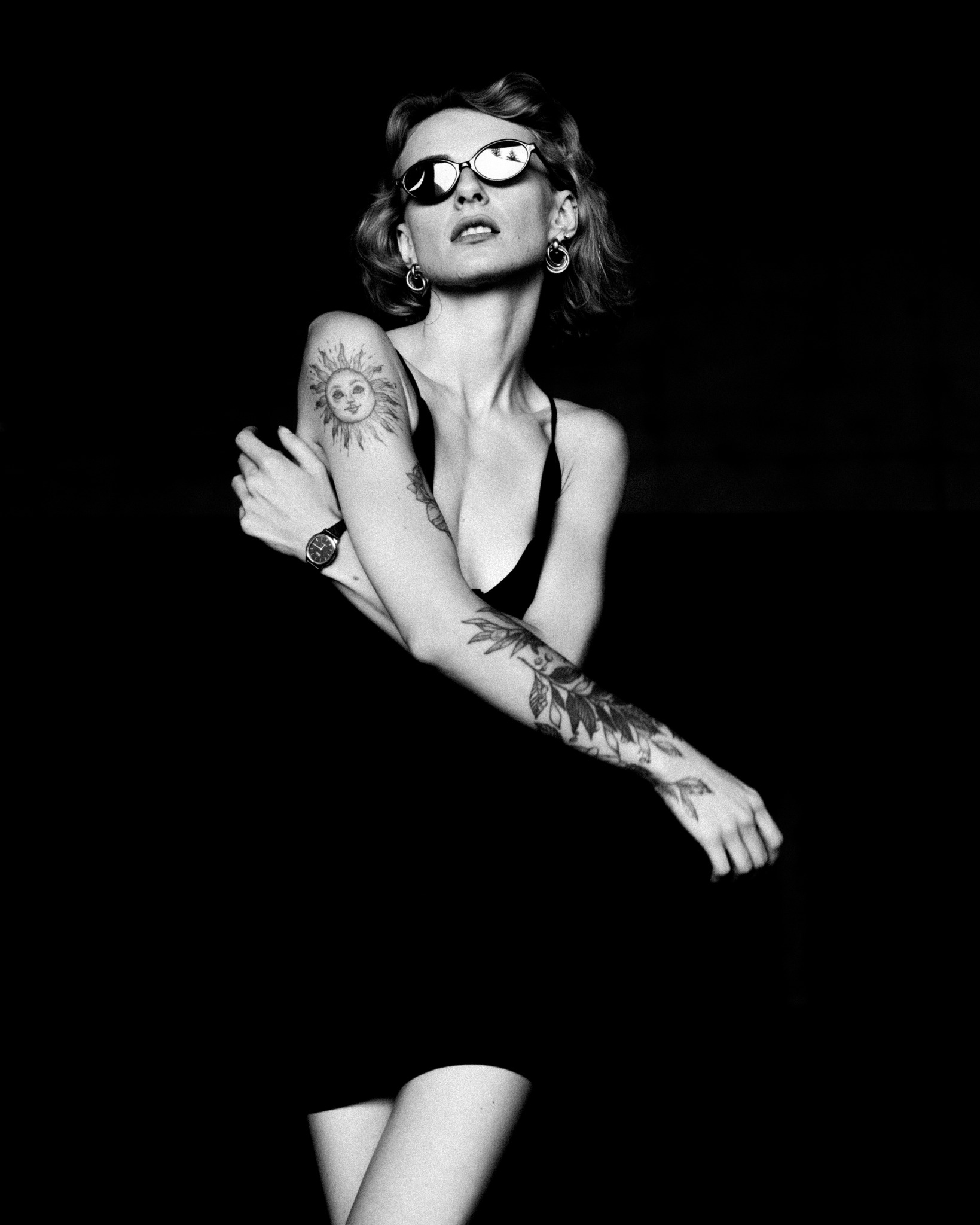 Czarno-biały portret kobiety w okularach, z tatuażami na ramionach, na ciemnym tle. Ekspresyjna poza i ostre światło.