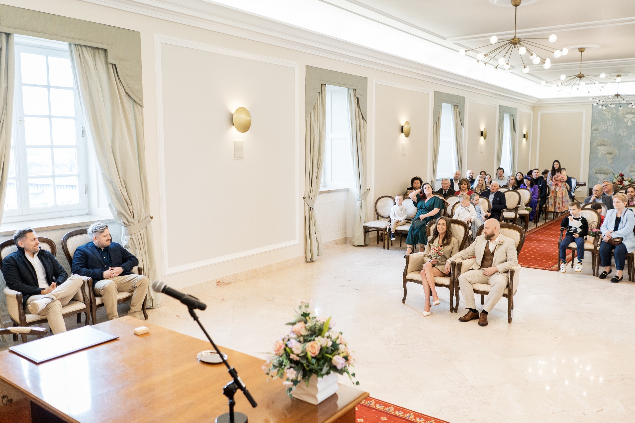 Elegancka sala ślubów: para młoda siedzi na krzesłach przed gośćmi, na pierwszym planie stół z mikrofonem i kwiatami, w tle okna z kremowymi zasłonami.
