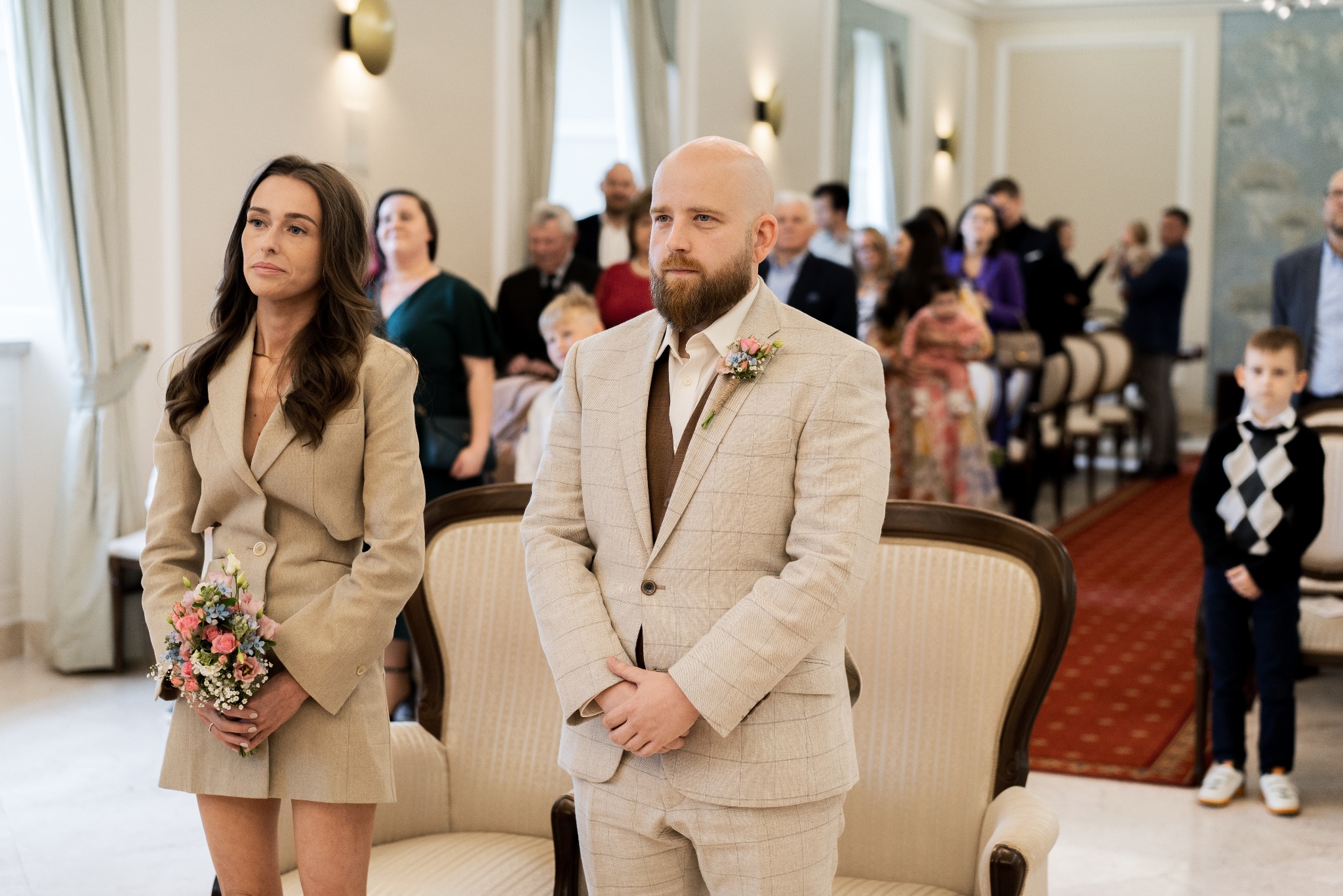 Para młoda w beżowych strojach podczas ceremonii ślubnej. Panna młoda trzyma bukiet, a pan młody ma butonierkę. W tle goście i dekoracje sali.