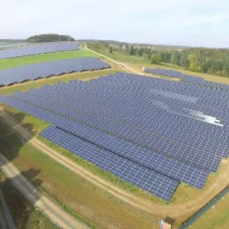 Rozległa farma fotowoltaiczna z panelami ustawionymi rzędami na zielonym polu, widok z góry, w tle las i błękitne niebo.