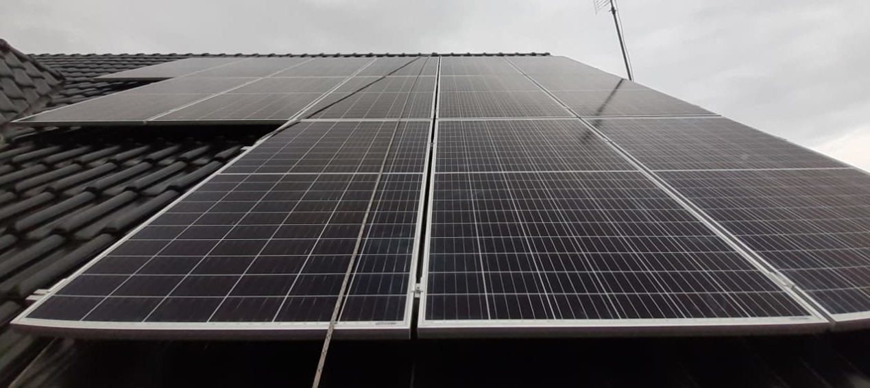 Panel słoneczny na ciemnym dachu z anteną w tle, pochmurne niebo.
