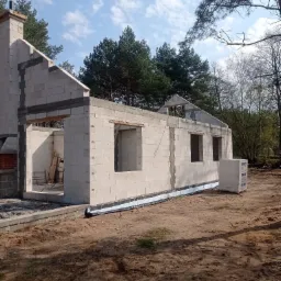 Surowy stan domu z białych bloczk&oacute;w, widoczne niezadaszone okna, komin, fundamenty z izolacją i paleta bloczk&oacute;w Solbet, otoczenie leśne.