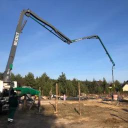 Betonomieszarka z wysięgnikiem pompuje beton na placu budowy fundamentów domu jednorodzinnego, widoczne szalunki i zbrojenia, w tle drzewa i domy.