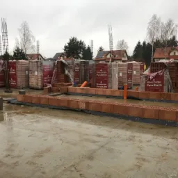 Wznoszenie ścian z pustaków ceramicznych Porotherm na mokrym fundamencie, widoczne zbrojenia i palety z materiałem budowlanym w tle zabudowa jednorodzinna.