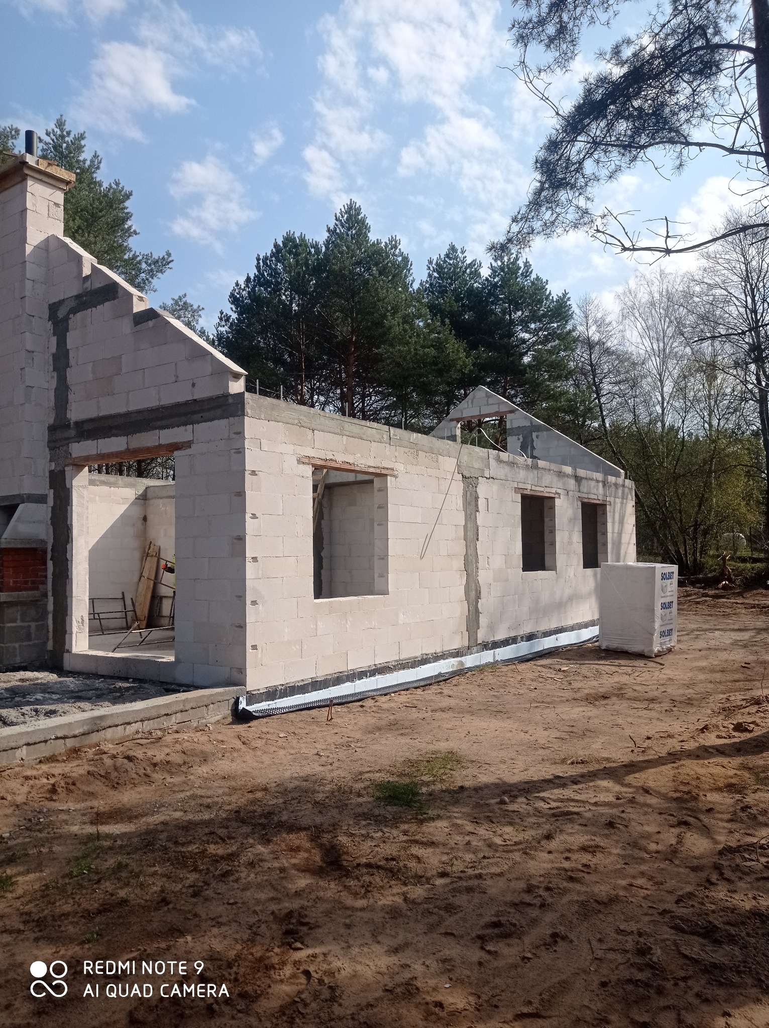 Surowy stan domu z białych bloczków, widoczne niezadaszone okna, komin, fundamenty z izolacją i paleta bloczków Solbet, otoczenie leśne.