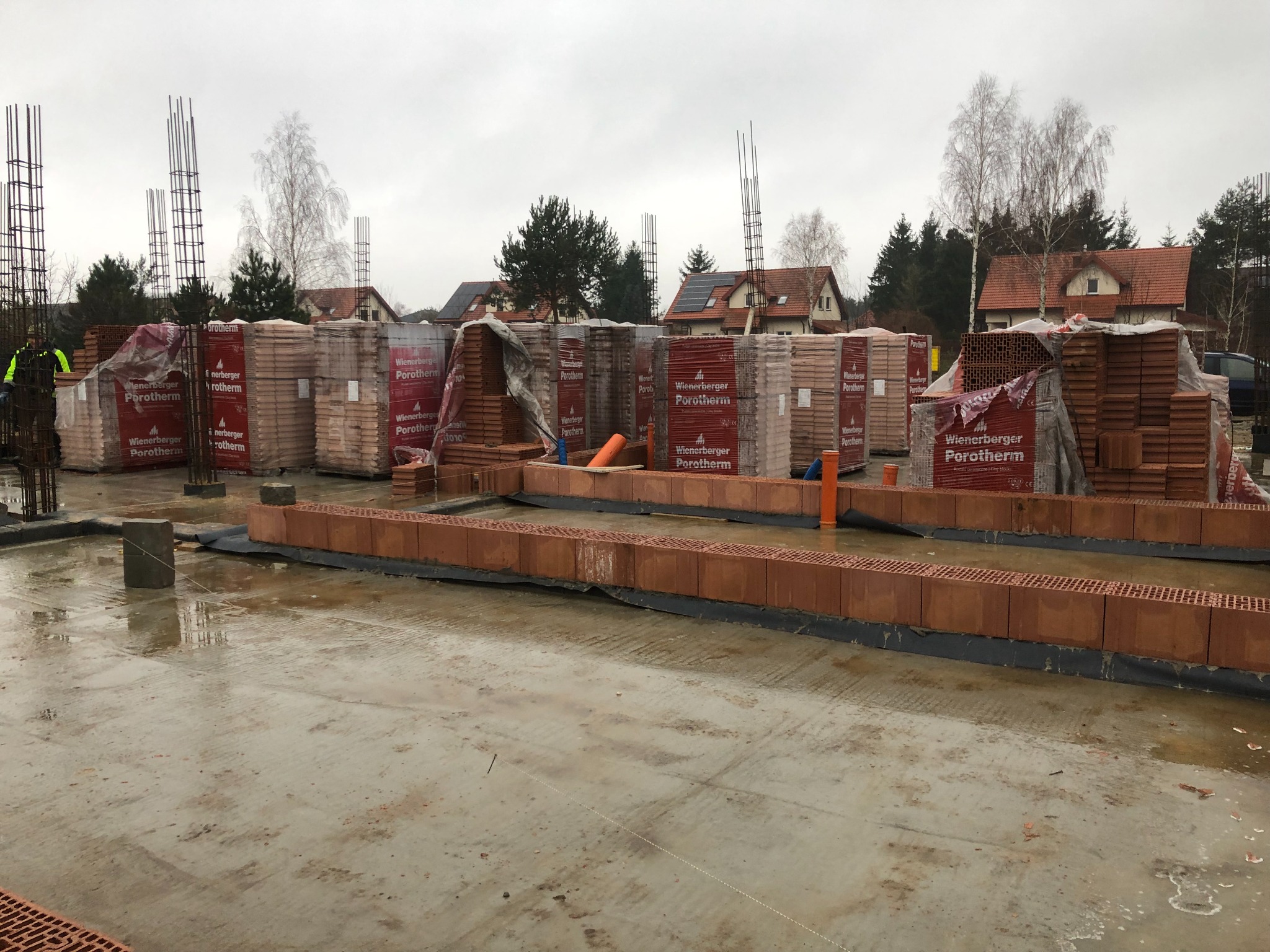 Budowa domu jednorodzinnego w Rajszewie: świeżo wylany fundament, częściowo wymurowane ściany z czerwonej cegły Porotherm, materiały budowlane w paletach z folią ochronną, wystające pręty...