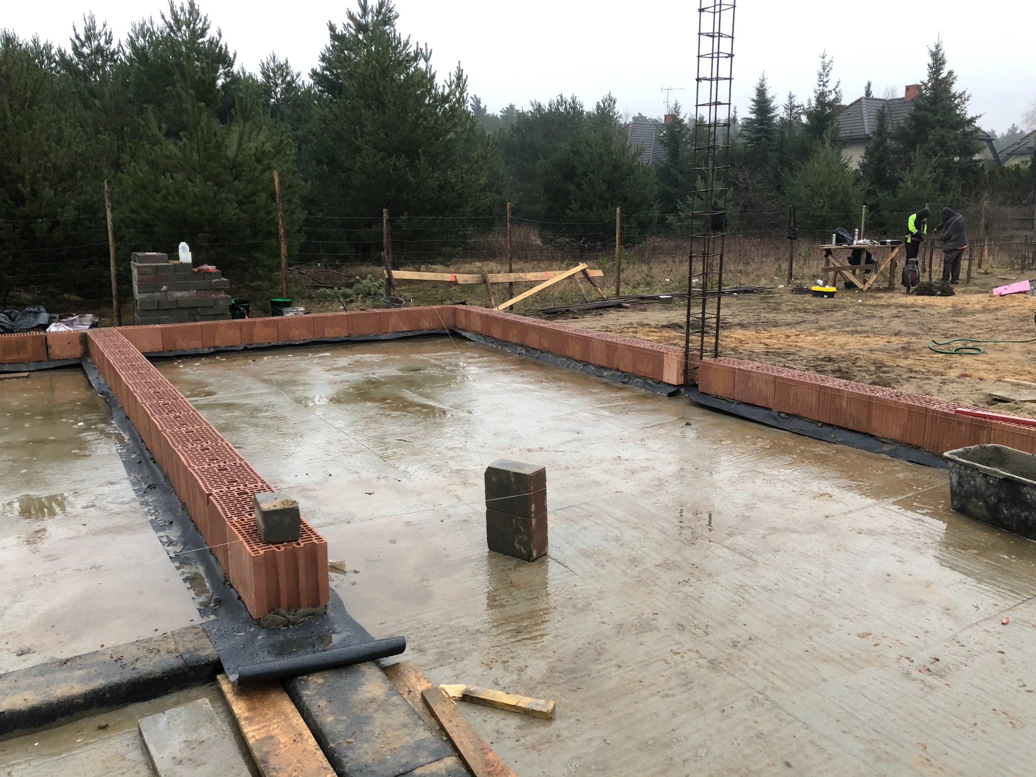 Wylany fundament pod dom z hydroizolacją i rozpoczętą budową ścian z pustaków ceramicznych; w tle las i pracownicy budowlani.