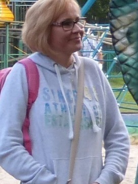 Kobieta w okularach, z plecakiem, w bluzie z kapturem, na tle placu zabaw.