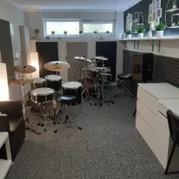 Wnętrze studia muzycznego z zestawem perkusyjnym, wygłuszonymi ścianami, biurkiem i krzesłem oraz roślinami w doniczkach na półce pod oknem.