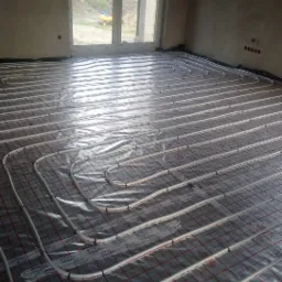 Równomiernie ułożone białe rury ogrzewania podłogowego na folii z czerwoną kratką w surowym wnętrzu z dużym oknem.