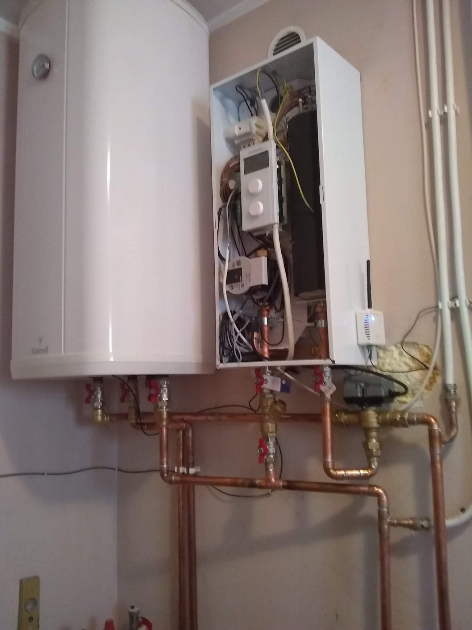 Biały zasobnik ciepłej wody i otwarty panel sterowania z widocznymi przewodami i elementami instalacji, połączone miedzianymi rurami z zaworami.