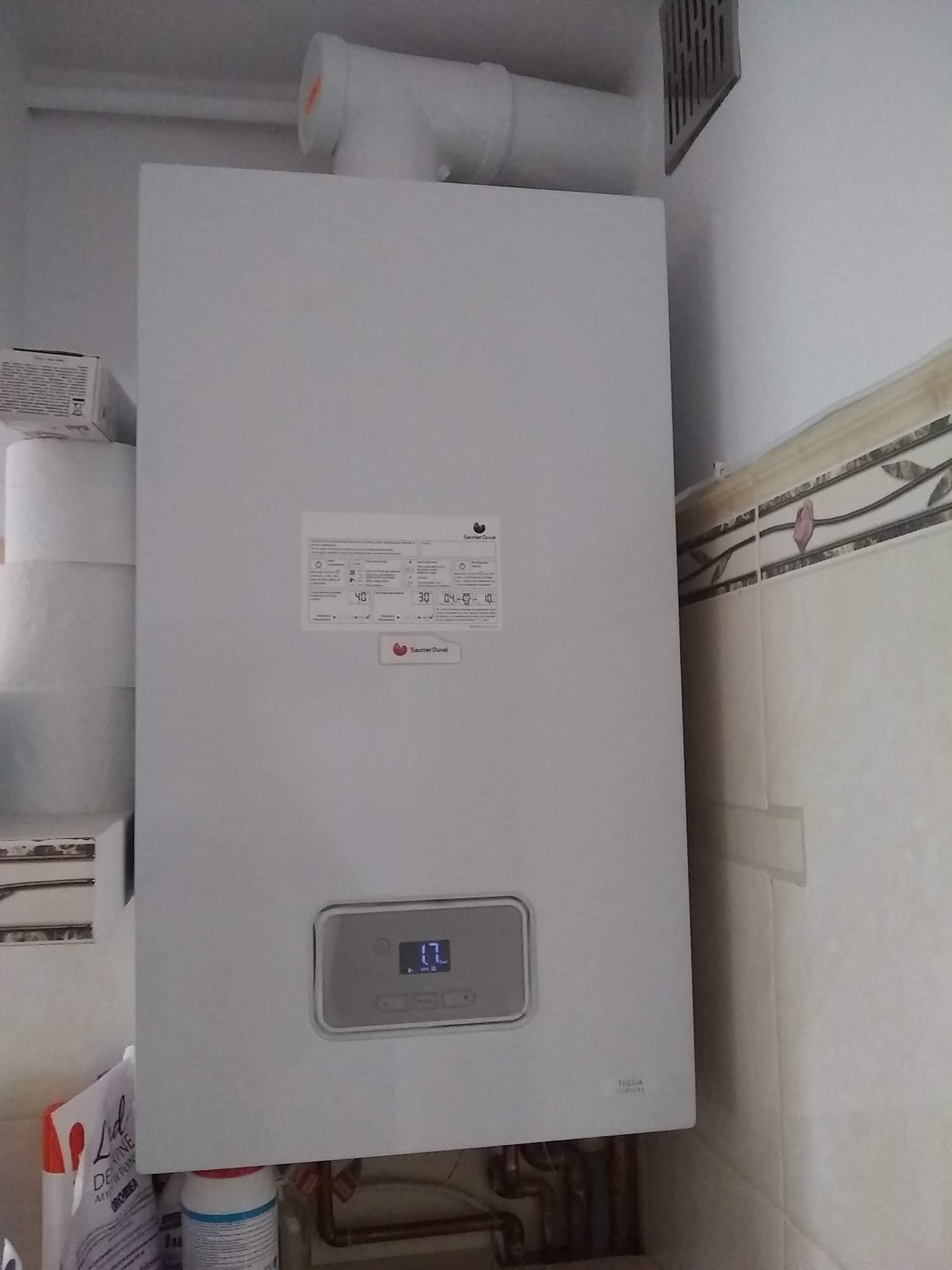 Biały piec gazowy marki Saunier Duval zamontowany na ścianie w pomieszczeniu z widocznymi rurami i elementami instalacji, obok rolki papieru toaletowego.