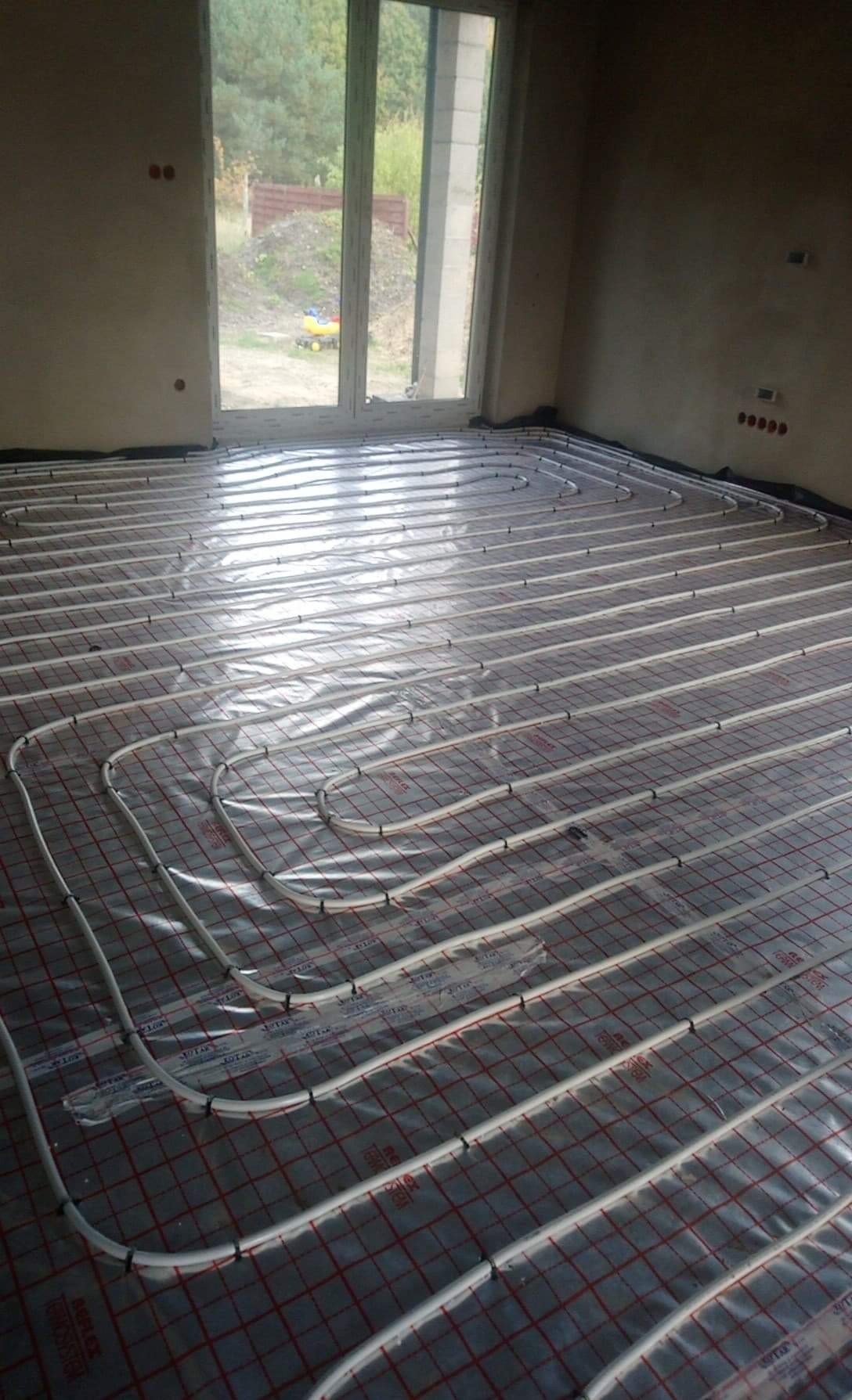 Równomiernie ułożone białe rury ogrzewania podłogowego na folii z czerwoną kratką w surowym wnętrzu z dużym oknem.