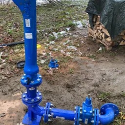 Niebieski hydrant przeciwpożarowy z zaworami i przyłączami kołnierzowymi, częściowo zmontowany na błotnistym gruncie, w tle stos drewna okryty plandeką i zalesiony teren.