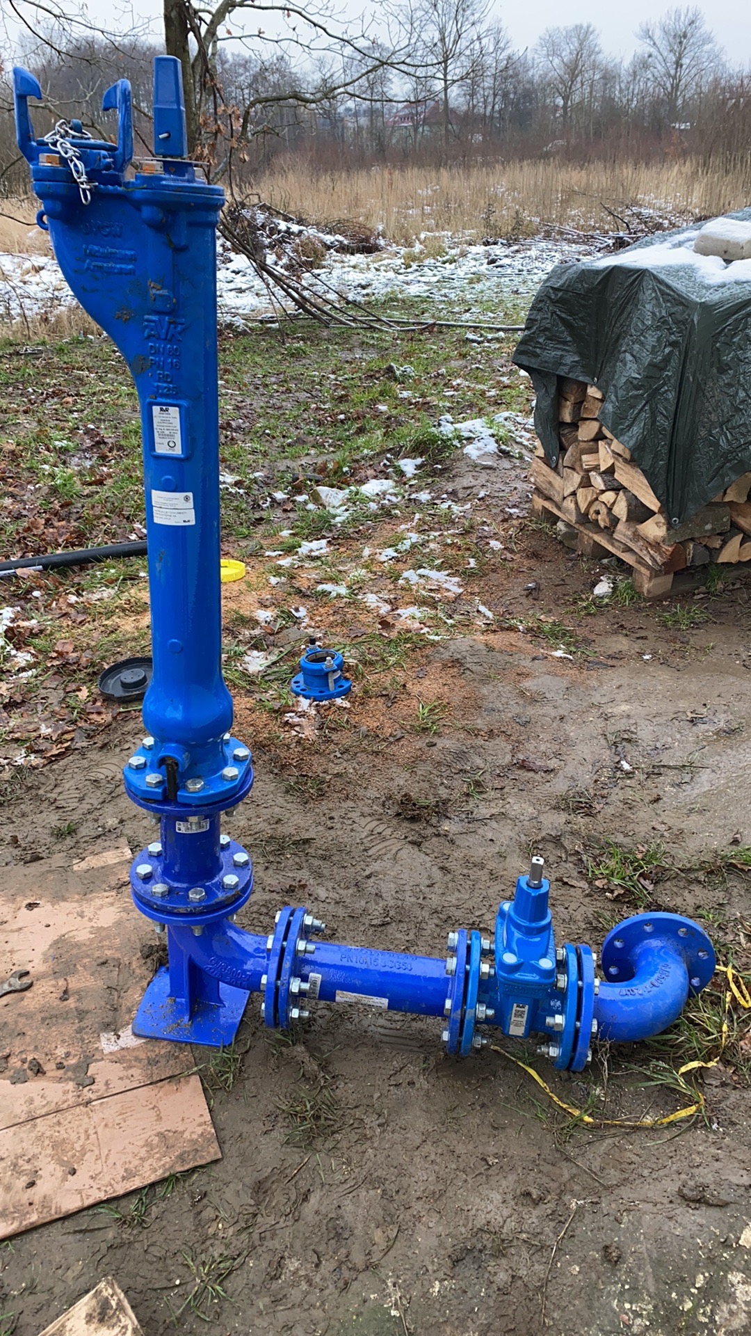 Niebieski hydrant przeciwpożarowy z zaworami i przyłączami kołnierzowymi, częściowo zmontowany na błotnistym gruncie, w tle stos drewna okryty plandeką i zalesiony teren.