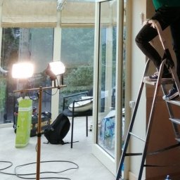 Projekt Mateusz Marszałek - Pracownik na drabinie podczas przygotowywania sufitu do prac wykończeniowych w jasnej oranżerii, widoczne oświetlenie robocze i narzędzia.