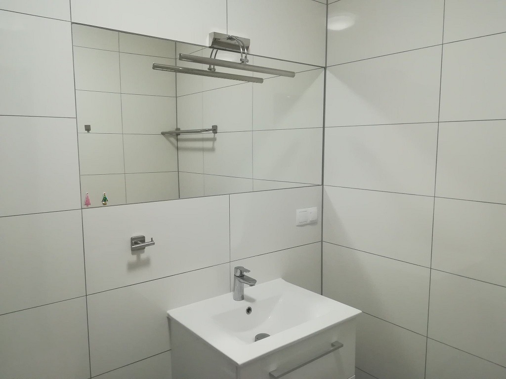 Biała łazienka z umywalką i dużym lustrem, na którym widać odbicie wieszaka na ręczniki, a na ścianie obok lustra przyklejone są małe naklejki choinek.