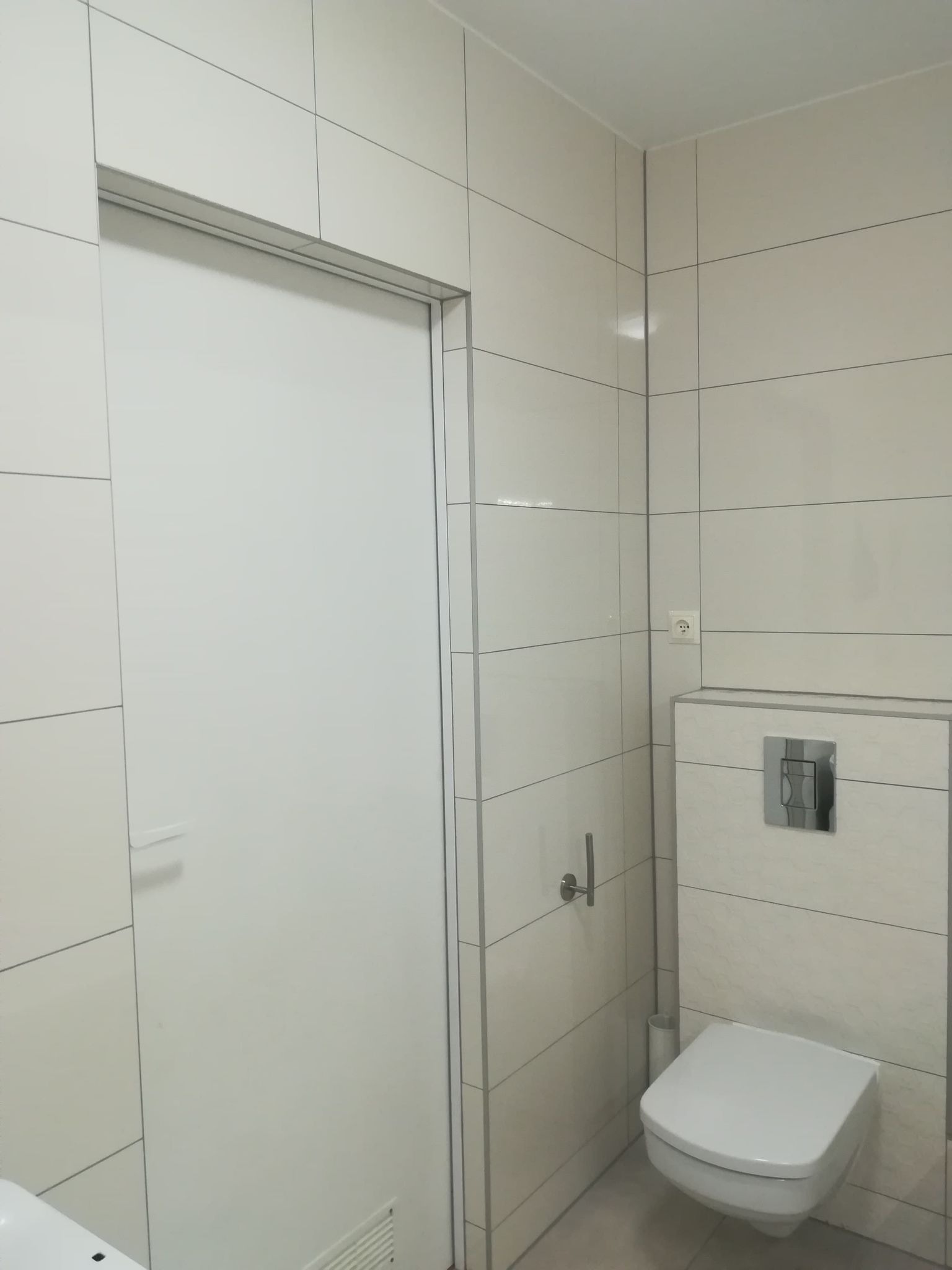 Wykończona łazienka z białymi, prostokątnymi płytkami na ścianach, wisząca toaleta z chromowanym przyciskiem spłukiwania i białe drzwi z aluminiową ościeżnicą.