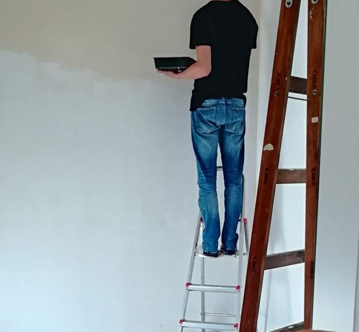 Mężczyzna w Gdyni maluje ścianę na biało, stojąc na drabinie, z widoczną kuwetą z farbą.