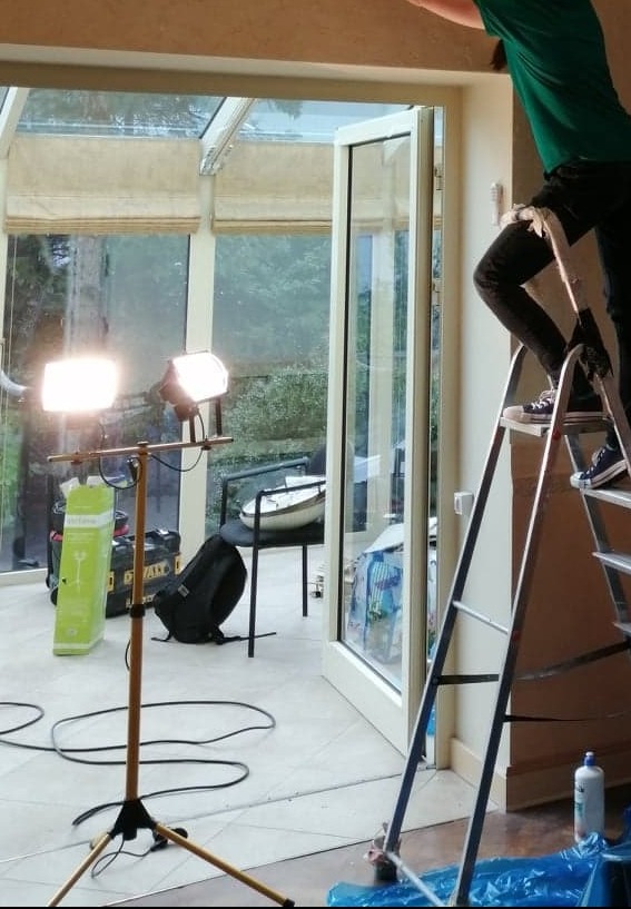 Pracownik na drabinie podczas przygotowywania sufitu do prac wykończeniowych w jasnej oranżerii, widoczne oświetlenie robocze i narzędzia.