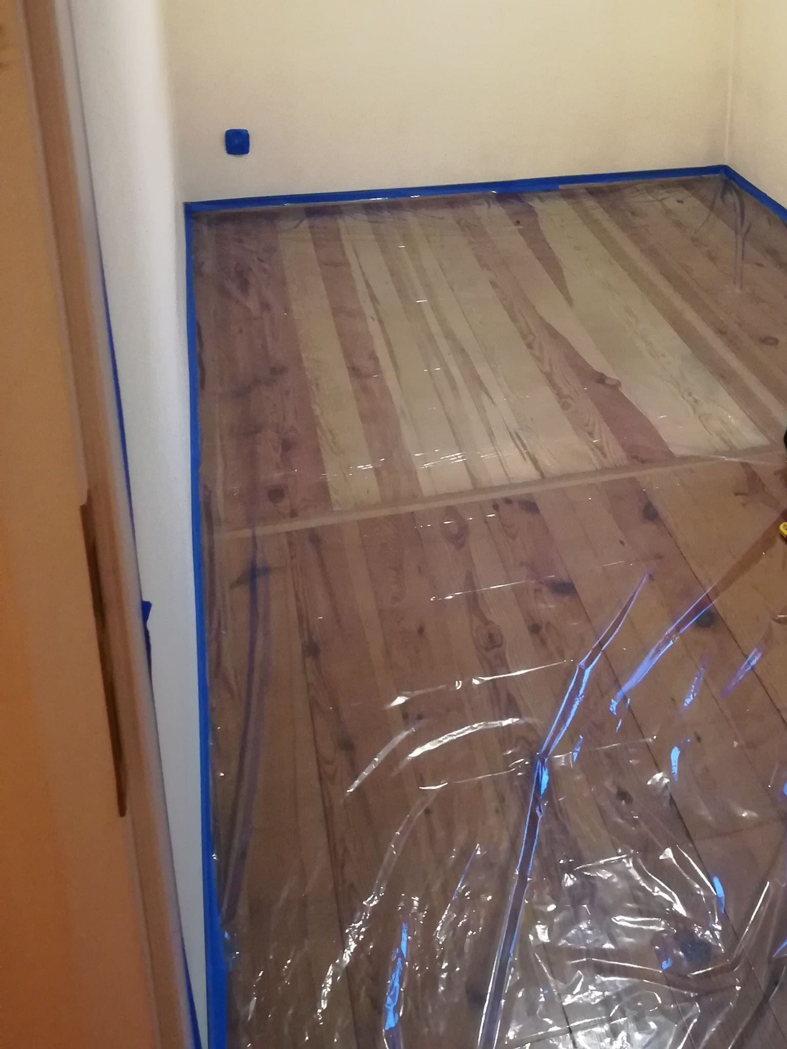 Przygotowanie do prac malarskich: zabezpieczona folią podłoga z drewnianych desek i niebieska taśma malarska przy ścianach i drzwiach.