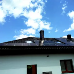 Czarny dach z dachówki ceramicznej z panelami słonecznymi i dwoma oknami dachowymi, kominy w kolorze dachu, biała ściana budynku, błękitne niebo z chmurami w tle.