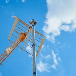 Antena telewizyjna typu Yagi-Uda na metalowym maszcie, zamocowana niebieską taśmą izolacyjną, na tle błękitnego nieba z białymi obłokami, ujęcie z dołu.