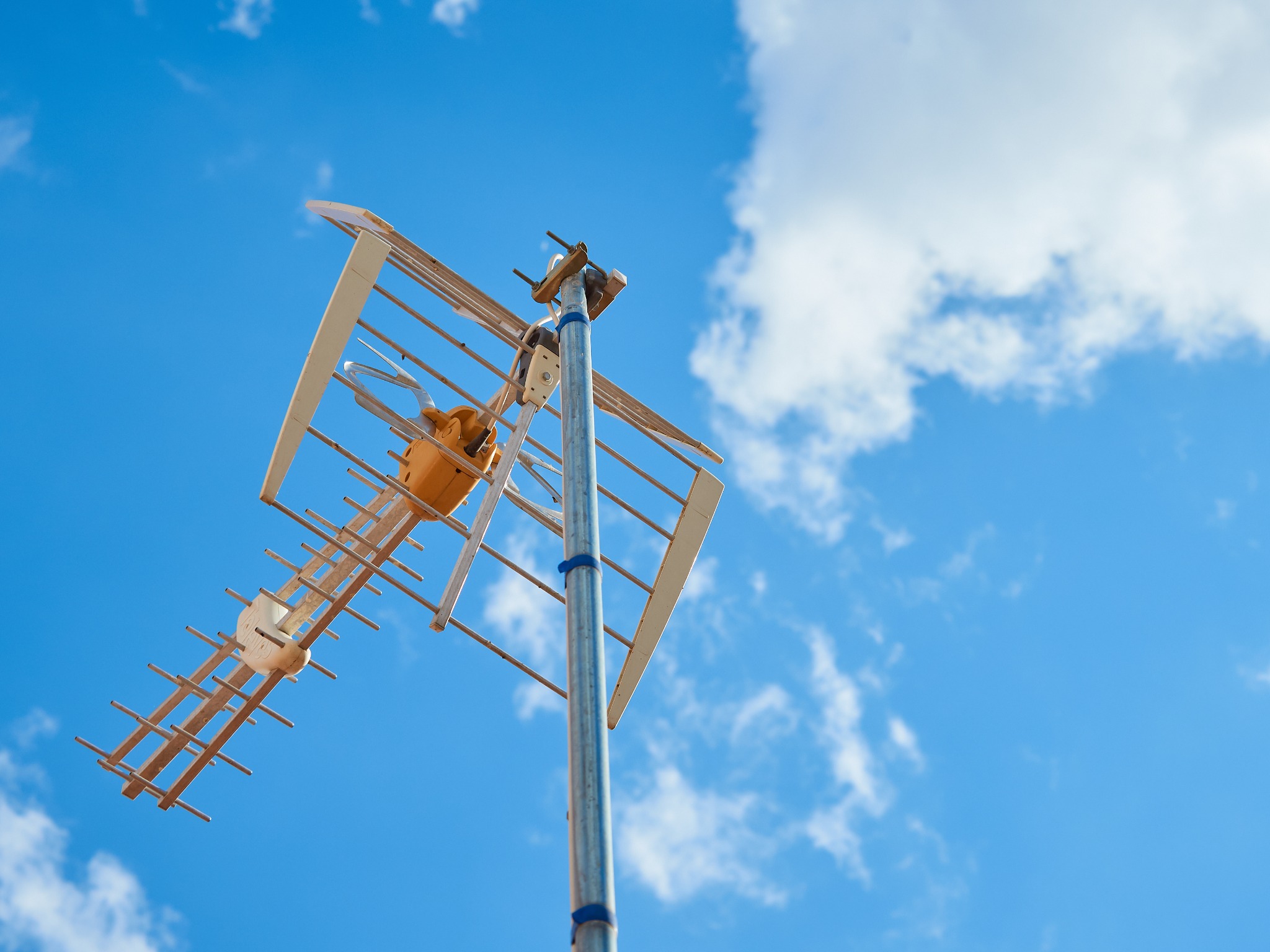 Antena telewizyjna typu Yagi-Uda na metalowym maszcie, zamocowana niebieską taśmą izolacyjną, na tle błękitnego nieba z białymi obłokami, ujęcie z dołu.