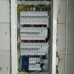 Rozdzielnica elektryczna z zainstalowanymi bezpiecznikami i przewodami w kolorach ż&oacute;łtym, zielonym, niebieskim i brązowym, zamontowana na surowej ścianie z betonu kom&oacute;rkowego.