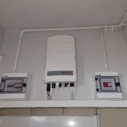 Biały inwerter SolarEdge zamontowany na ścianie między dwiema białymi skrzynkami elektrycznymi. Widoczne kable i przewody prowadzone w białych rurkach na ścianie. Biała ściana i sufit. Czujnik...