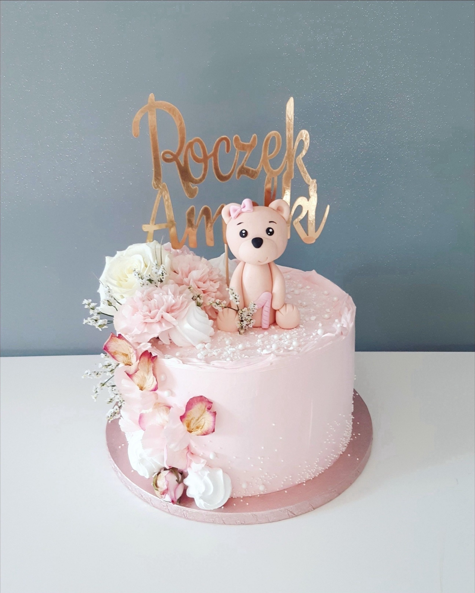 Tort na roczek w pastelowych odcieniach różu, zdobiony kwiatami, figurką misia z cyfrą '1' i złotym napisem 'Roczek Amelki'.