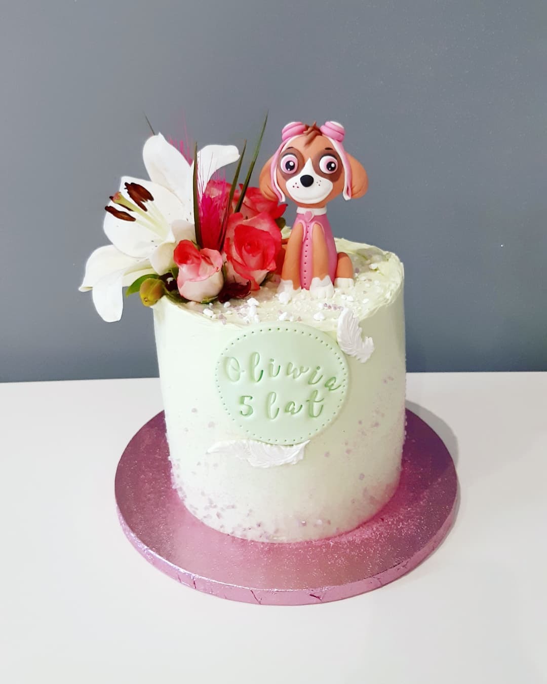 Tort urodzinowy z dekoracją kwiatową i figurką pieska Skye z Psiego Patrolu, na różowym podkładzie, z napisem 'Oliwia 5 lat' w zielonym kółku.