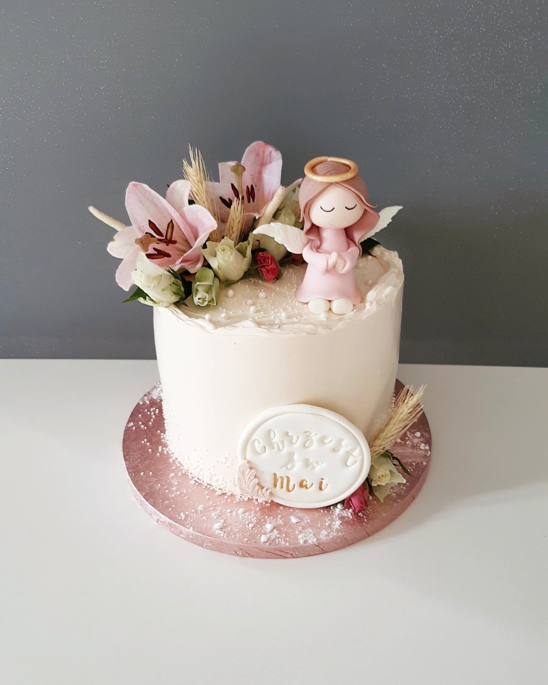 Tort z kremem, dekorowany figurką aniołka z aureolą, liliami, różami i napisem 'Chrzest Św. Mai' na białym tle.