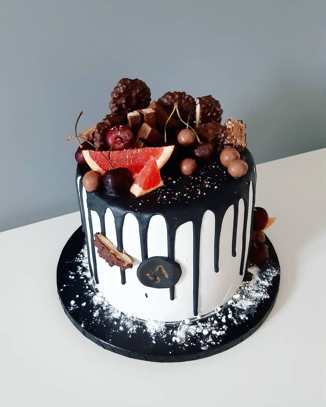 Tort urodzinowy z białym kremem, czarną polewą, dekorowany czekoladkami, wiśniami i grejpfrutem, z numerem 31 na czarnym znaczku.