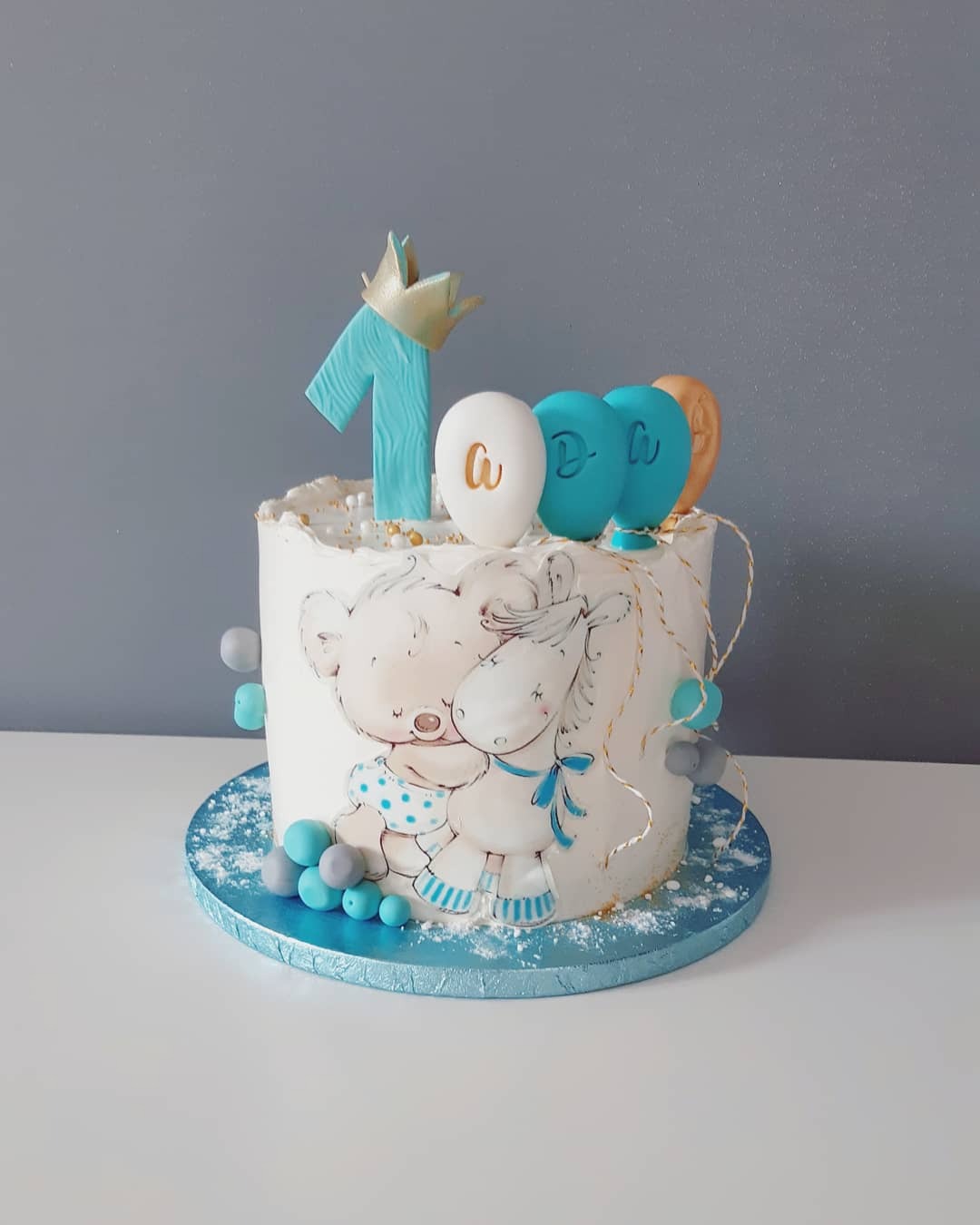 Tort urodzinowy dla roczniaka z ręcznie malowanymi postaciami misia i jednorożca, dekorowany liczbą '1' z koroną i balonami w odcieniach błękitu i złota.