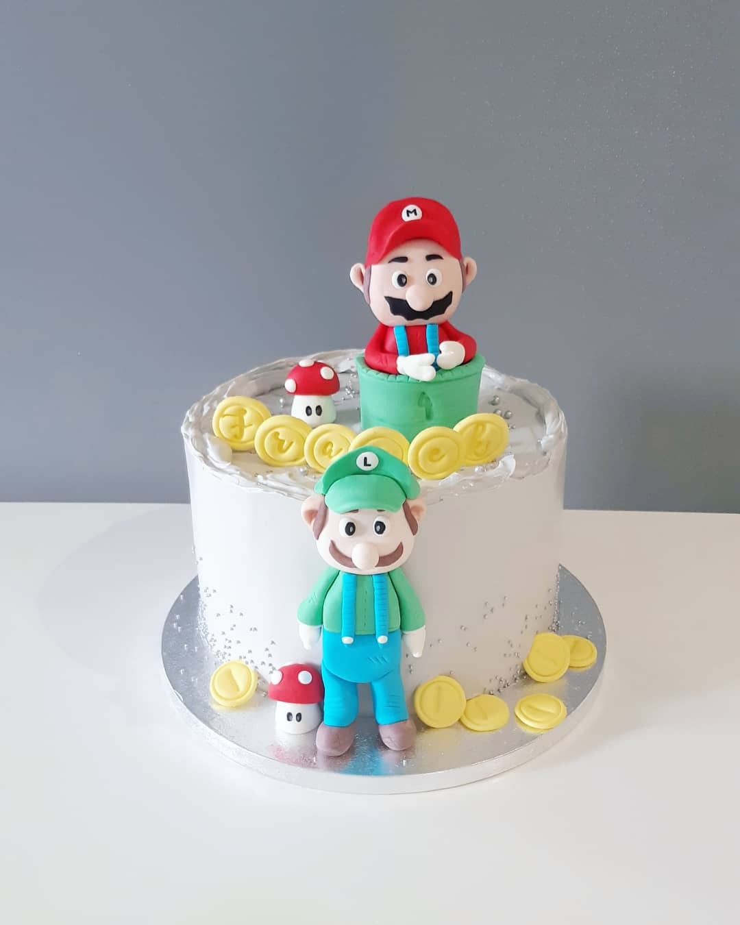 Tort urodzinowy z białym kremem, dekorowany figurkami Mario i Luigiego z masy cukrowej, grzybkami i złotymi monetami.