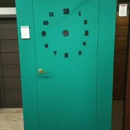 Turkusowe drzwi wewnętrzne z czarnymi cyframi tworzącymi zegar, złota klamka po lewej stronie, widoczne inne modele drzwi w tle.