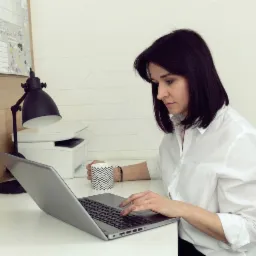 Kobieta w białej koszuli skupiona na pracy przy laptopie, z kubkiem w zygzaki obok, biurko z lampką, drukarką i kalendarzem w tle.